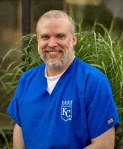 Smiling man with a beard wearing a blue medical scrub with a "KC" logo.