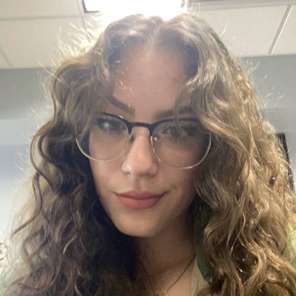 Young woman with curly hair wearing glasses and smiling slightly.