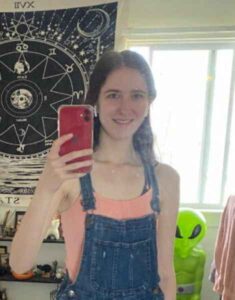Young woman taking a selfie, wearing overalls and standing before a tapestry.