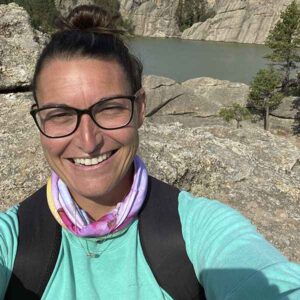 Woman smiling in a selfie with rocky terrain and pine trees near a river in the background.
