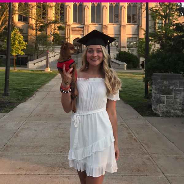 Graduate in white dress holding a small dog in front of a building.