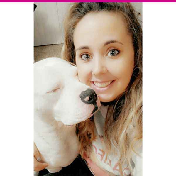 Woman smiling with a white dog nuzzling against her face.